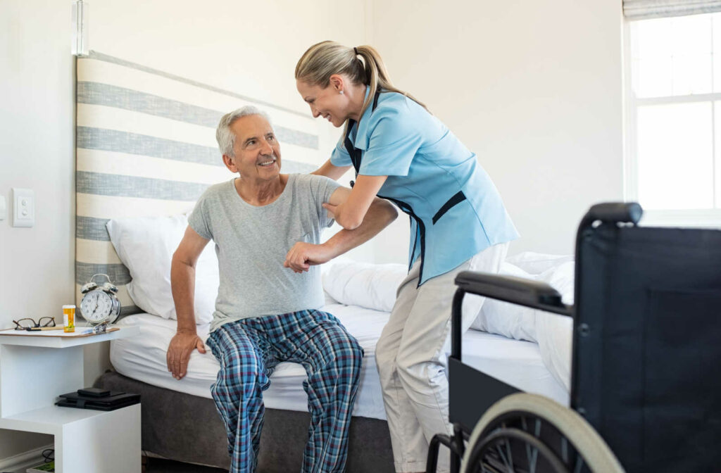 A female respite care professional is assisting a senior male going up from his bed going to his wheelchair.